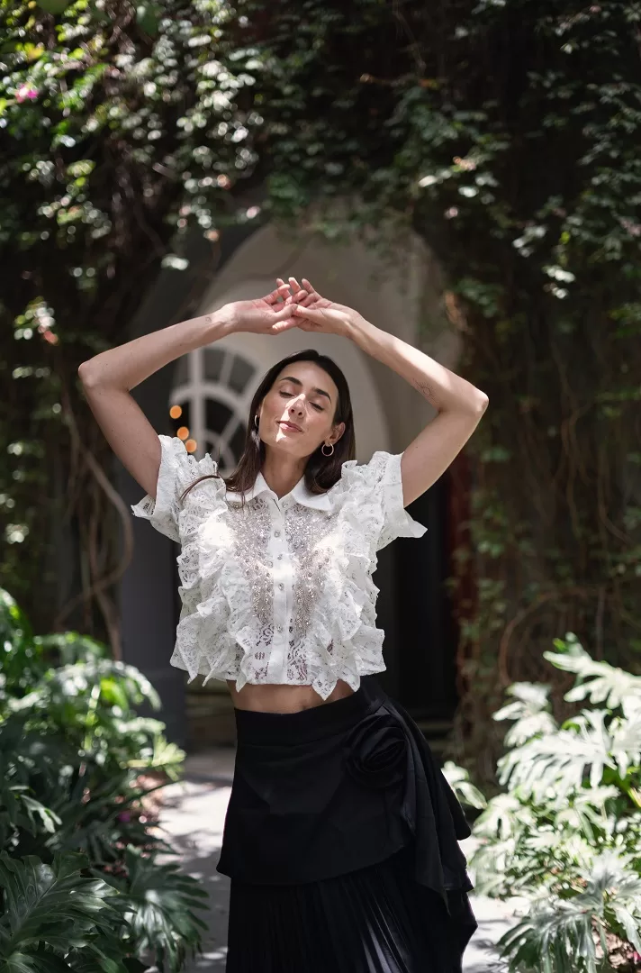Camisa de encaje con volantes, corte cropped y bordado de pedrería en forma de alas. Disponible en blanco y negro. Elegancia con actitud para tus ocasiones más especiales.