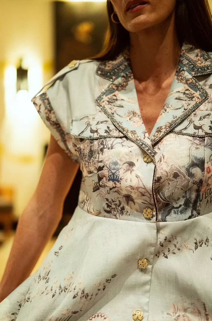 Vestido camisero con print artístico estilo toile de Jouy en tonos suaves, cuello estructurado, hombreras con botón dorado y pedrería en solapas. Elegancia atemporal con espíritu moderno.
